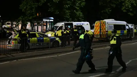 Police walking down the road in Bristol in riot gear. In the back of the image two police vans and a police car are parked in the road.