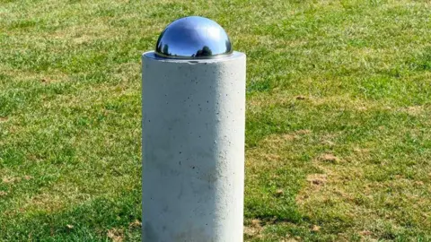 A cylindrical concrete pillar standing on a paved base in a grassy field, topped with a small reflective metal sphere representing a planet marker in the Otford solar system model.
