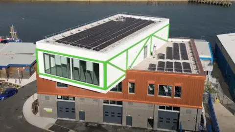 An aerial view of the Energy Central Learning Hub in the Port of Blyth which is a white, green and brown building in front of the North Sea. The top part of the building is jutting out to the side. 