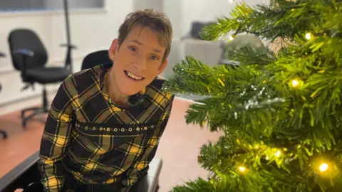 A woman in a yellow dress sits in a wheelchair smiling next to a sparkly Christmas tree