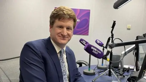 A man in a blue suit sat next to a purple microphone with write writing reading "BBC Radio Kent".