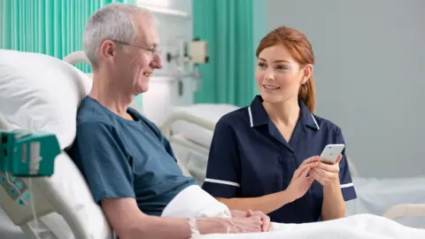 A man, in a hospital bed, wearing a blue top, with short grey hair and glasses, smiling, with a nurse, in a blue uniform, with long red hair, tied back, smiling at him, while holding a phone. There is a bed behind them, all with white sheets on. 