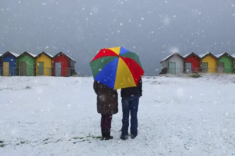 Alison Leddy A couple holding an umbrella standing in snow with beach huts in the background