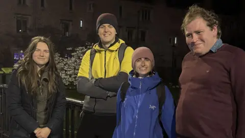 Two men and two women on Magdalene Bridge, Cambridge, simling. It is dark. The two women and the man standing in between them are wearing walking jackets. The man on the right is wearing a jumper over a shirt. 