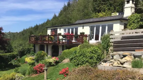 Photograph pf the remote bungalow in Maenan near Llanrwst, overlooking the Conwy Valley. The white building has a wooden patio perched above a sloping flower filled garden.