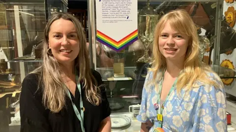 BBC/Seb Cheer Two women with blonde hair look into the camera, smiling. One is wearing a black top, the other's is light blue and floral. They are both wearing turquoise lanyards, and are standing in front of a white board with rainbow trim, next to a