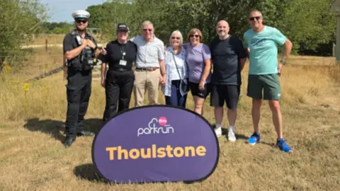 Wiltshire Police PC Slade, PC Howick, David Painter and his wife Jackie, and other parkrun volunteers standing in a row with arms interlinked at Thoulstone Park with dried-out grass in the foreground and trees in the background.