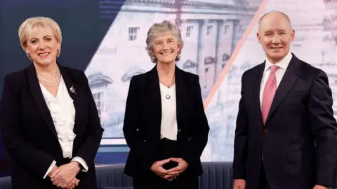 Conor O'Mearain/PA Wire Heather Humphreys, Catherine Connolly and Jim Gavin pose and smile for a photo in a TV studio. Humphreys and Connolly are both wearing black suits over white tops. Gavin is wearing a black suit with a white shirt and a pink tie. 
