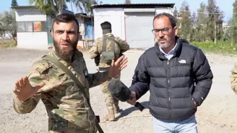 The soldier of the Ministry of Defense of Syrian, Mahmoud Al-Haik, is gestured as the BBC News Arabic correspondent, Ferras Kilni, has a microphone to Al-Haik