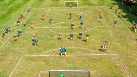 Timothy Welstead A wide shot of a football match scene in a model village. Players in red and blue strips can be seen dotted around the pitch. At the far end, the streaker can be seen invading the pitch.