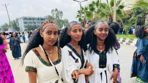 Girmay Gebru/BBC Three girls wearing white and black dresses with their hair in braids pose and smile for the camera. They are outside and there are some palm trees in the background and other women wearing dresses who are participating in the festival in Mekelle, Ethiopia. Sunday 24 August 2025.