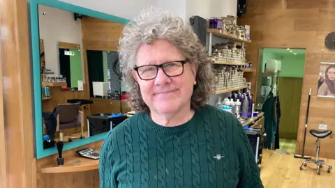 A man stands in a hair salon. He has lots of curly grey hair and is wearing a green Gant jumper. He has large black-rimmed glasses on.