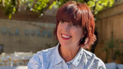 Catherine Warrilow Catherine Warrilow smiles in a garden. She has red hair, nose jewellery and wears a blue top.