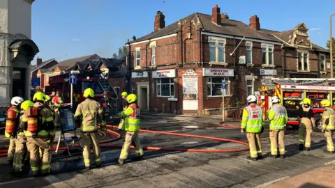 About 10 firefighters wearing light brown uniforms and high-vis jackets and helmets, some with breathing apparatus, stand in a street with two fire engines and red hoses. In the background, smoke can be seen rising into a blue sky from a single-story building with damaged and blackened roof gables. The damaged building is behind a grander, red-brick, two-story building which has signs reading "Swinton WMC".