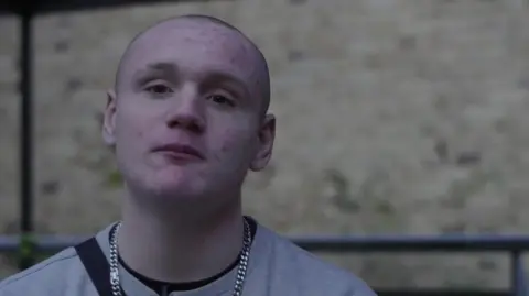 Teenage boy in a grey Champion sweatshirt with silver chain looks at the camera