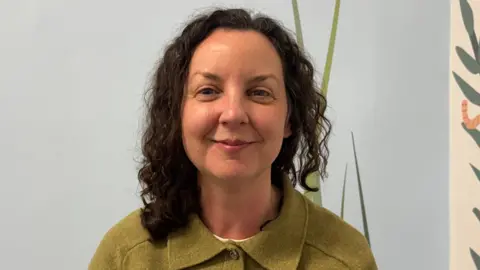 Cathy - dark curly hair wearing a green buttoned top, standing against a light -coloured wall with stylised leaf artwork visible to the side.

