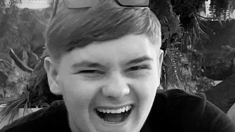 Harvey Willgoose, a smiling teenage boy wearing a dark top and glasses on his head, in a monochrome photograph.