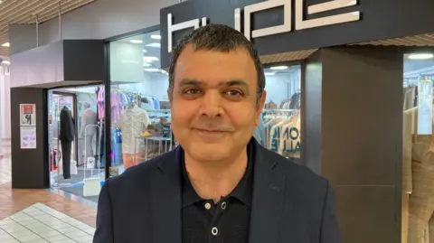 A smiling man with dark hair and wearing a blue suit jacket and black polo shirt and shop sign behind him spelling out the name HYPE