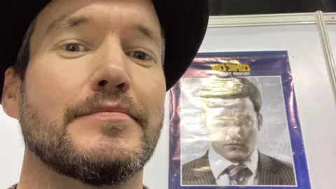 Gareth David-Lloyd Gareth David-Lloyd wearing a black hat, stood in front of a poster of his younger self in a pin-striped suit