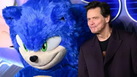 Getty Images Uma foto tirada na estreia de um filme mostra Jim Carrey parado ao lado de uma pessoa com uma fantasia azul gigante e fofa de Sonic the Hedgehog.