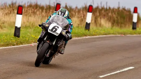 CLASSIC TT Michael Dunlop riding over the mountain on a black classic bike with the number six on the front. His leathers are coloured black, purple and turquoise.