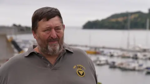 A picture of Dr Harry Bennett. He has a short grey beard and is wearing a grey polo shirt. He has black hair.