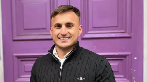 A man with short dark hair and a dark top smiles at the camera, with a purple door behind him