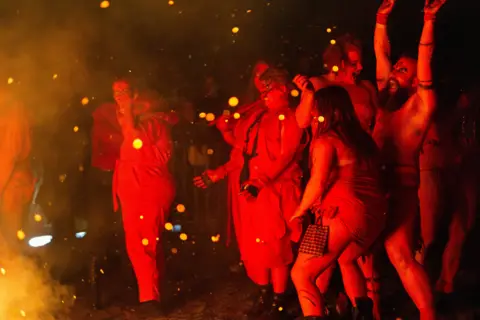 Zhi Kang Chua A group of people are covered in the red glow from the fire. They are all dancing a embers fly around them.