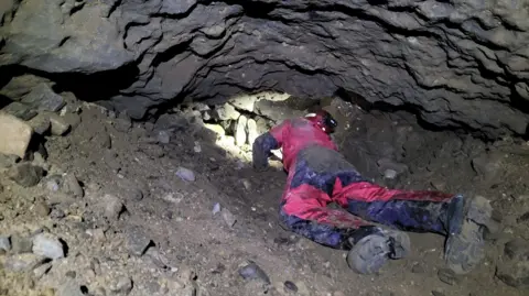 Assynt Mountain Rescue Team A rescue volunteer dressed in red and black clothing follows another volunteer in a confined, rocky space.