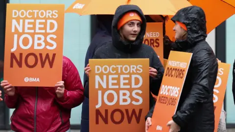 Several doctors on strike, wearing orange BMA hats and hooded raincoats and holding up placards calling for more pay, saying 'patients need doctors, doctors need jobs'' outside University Hospital Liverpool on Friday.