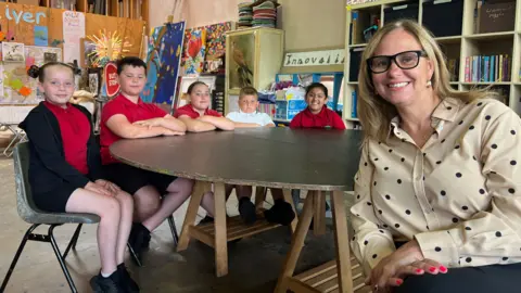 Helen Godwin, the West of England Mayor, is seated smiling with a group of schoolchildren in a brightly lit classroom. The children are gathered around a table, some leaning in with interest. The setting reflects a typical primary school environment, with colourful posters and school supplies in the background.