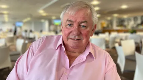 David Graveney. He has short grey hair and is wearing a pink shirt. He is looking at the camera and smiling