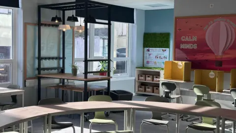 A classroom, it has different zones, in the front there are arched tables, behind there are higher tables with stools, and at the back right there are individual tables with screens for privacy, it looks modern and colourful.