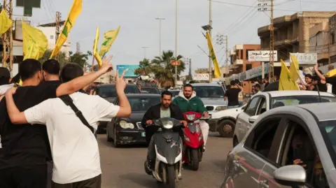 Two men on motorcycles ride among traffic, as people make victory signs and fly yellow Hezbollah flags, in Nabatieh, Lebanon (17/04/26)