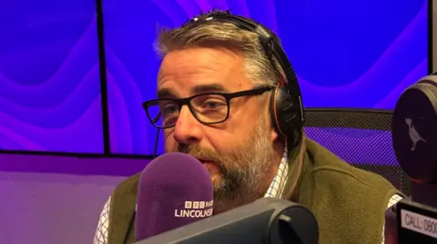 Richard Davies has grey hair and a grey and brown beard. He's wearing a green jacket and is sitting in front of a purple microphone. Behind him are an array of purple screens.