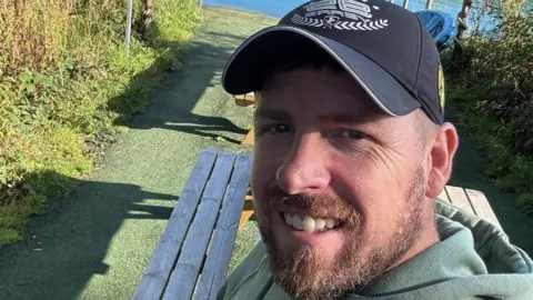 Chris Hughes A man with a hat, beard and green hooded top, takes a selfie while standing by a lake.
