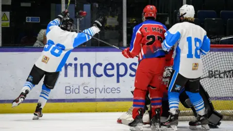 Paisley Pirates Three ice hockey players, two wearing white and blue kits while another wears a red and dark blue kit on an ice rink near the goals. 