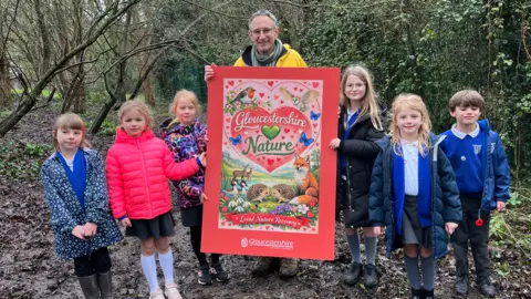Six young children in school uniform with coats on in a muddy wood with councillor Martin Horwood in the middle. Horwood is wearing a yellow coat and scarf and holding a bright pink Valentine's day card which is very large, covering his hole body. On it it says 'Gloucestershire nature', with a green heart in between. There are hearts all over the card as well as hedgehogs, a robin, deer, ducks and a barn owl in a nature-themed scene. At the bottom of the card it says 'local nature recovery', with the Gloucestershire County Council logo.