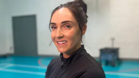 Aimee Oliver. She has dark hair tied back, wearing black gym clothing.