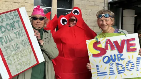 Three campaigners holding placards which say 'please don't pull the plug on Stroud lido' and 'Save our Lido'. The person in the middle is wearing a red lobster costume and the women either side of him are wearing swimming goggles.