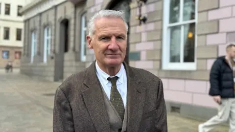 A man wearing a brown blazer, waistcoat, tie and white shirt. He is standing on a pavement outdoors next to a brick building. He is looking into the camera.