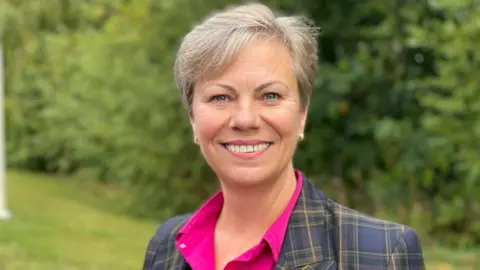 A smiling grey-haired woman standing on a lawn and in front of bushes, wearing a tartaned blazer and pink shirt.