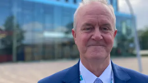 Man smiling at the camera wearing a blue suit jacket, white shirt and blue patterned tie. He has a North Northamptonshire Council lanyard round his neck. In the background is a large glass building.