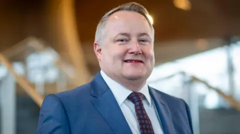 Getty Images A head and shoulders shot of Darren Millar, looking towards the camera wearing a burgundy and blue tie, a white shirt and a blue suit. The Senedd's wooden ceiling is in the background and out of focus.