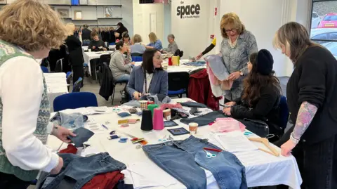 A group of women are using a variation of sewing materials to repair donated jeans. They are standing and sitting round the table talking to each other. 
