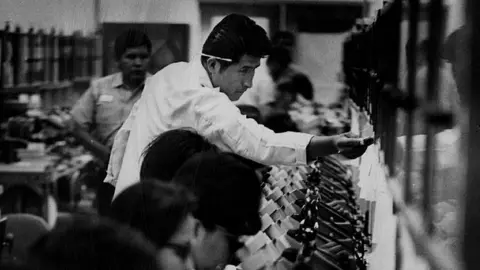 Getty Images A black and white picture of a row of workers looking into microscopes. There is a man in a white shirt standing who looks like he is adjusting something.