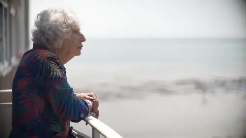 Carol Turansky standing on a balcony. She is wearing a leaf print dress and you can see her side profile. She has grey permed short hair and you can see the sea in the background. She is looking out to the horizon.