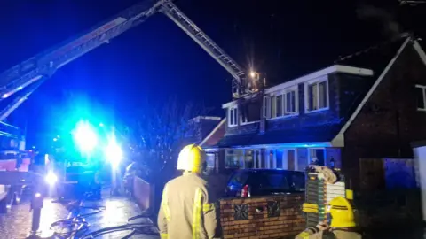 Two firemen in the foreground looking towards a house at night time. There is a crane with platform outside a burnt out window.