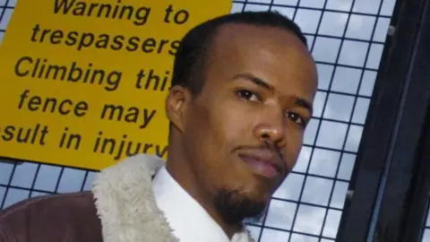 Newsquest/SWNS Close-up headshot of Essa Suleiman who has a small goatee beard and is wearing a sheepskin jacket, standing in front of a fence with a yellow sign saying "warning to trespassers"