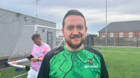 A smiling man is wearing a green and black "Just a Chat" football jersey stands on an outdoor football pitch. Another player in a pink shirt is looking down at his phone near a goal post under an overcast sky